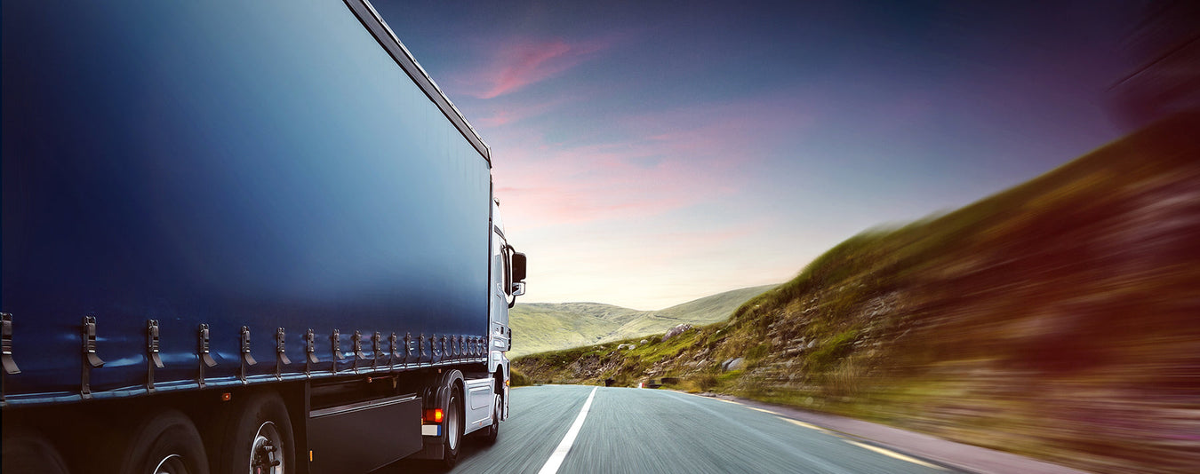 a scenic image of a navy lorry travelling down a main road underneath a pink and blue sky